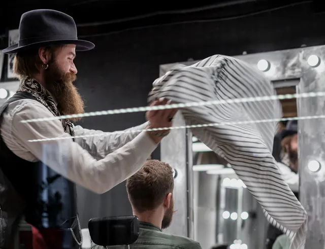 Barber shaking out a striped cape behind a seated client